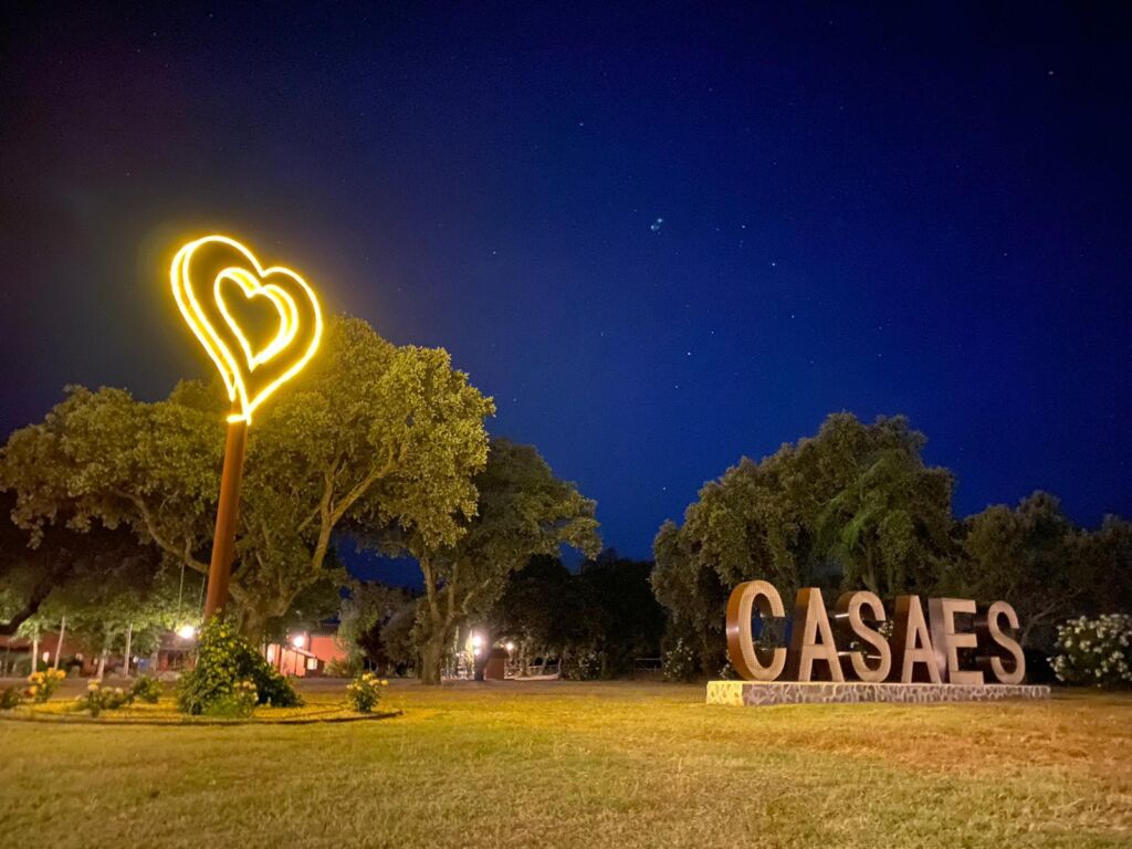 Un corazón de neón amarillo resplandeciente se alza sobre el césped junto a grandes letras metálicas que deletrean CASAES, rodeado de árboles e iluminado bajo un cielo nocturno oscuro con estrellas visibles.