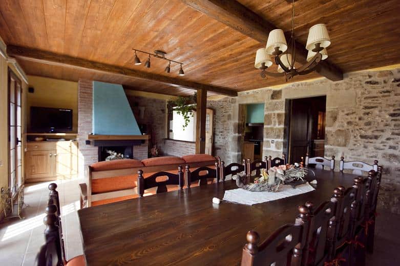 Un comedor rústico con una larga mesa de madera, sillas de madera oscura, una pared de piedra, una chimenea con una chimenea azul, una lámpara de araña y un acogedor sofá naranja al fondo.