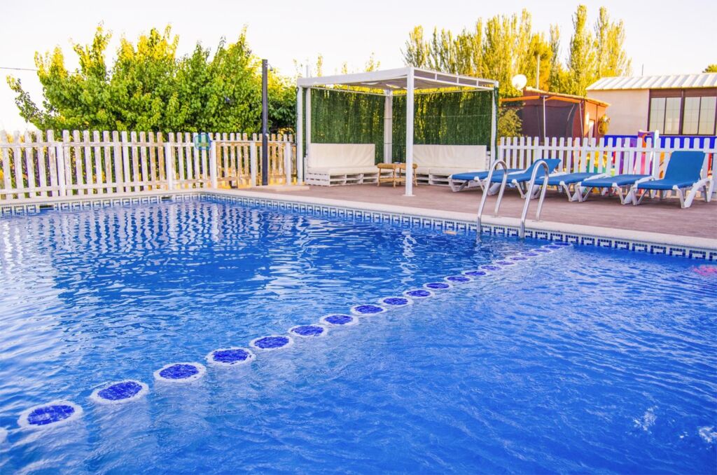 Una piscina al aire libre de color azul claro con una cuerda divisoria, rodeada de tumbonas, una zona de asientos a la sombra y una valla blanca, con árboles y edificios al fondo.