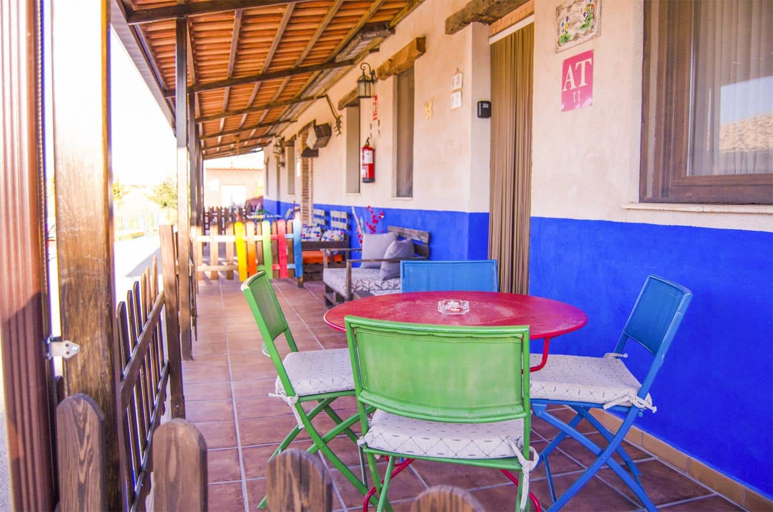 Un patio colorido con una mesa roja y sillas verdes y azules, colocadas sobre un suelo de baldosas. La pared exterior está pintada de azul y beige, con vigas de madera y diversos adornos. Contra la pared hay un banco acolchado.