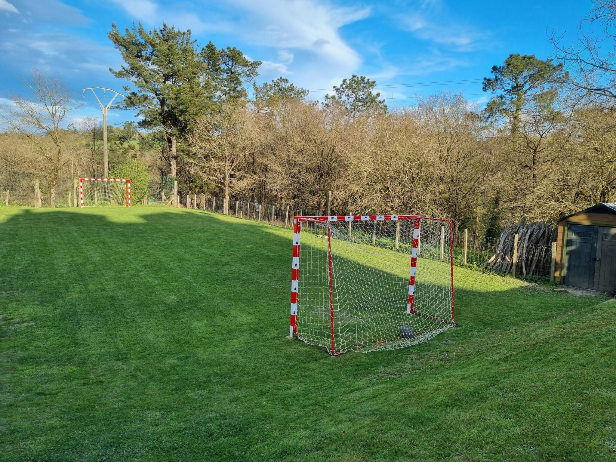 Un campo de fútbol de hierba al aire libre con dos porterías rojas y blancas, una cerca, en primer plano, con un balón de fútbol dentro, y la otra más alejada; árboles y cielo azul al fondo.