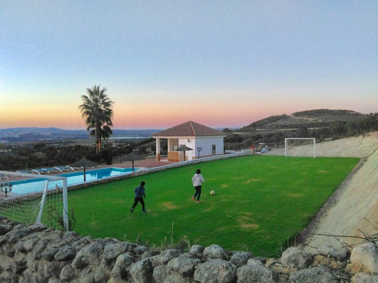 Dos niños juegan al fútbol en un pequeño campo de hierba delimitado por un muro de piedra, con una piscina, una palmera y una casa blanca al fondo, rodeados de colinas al atardecer.