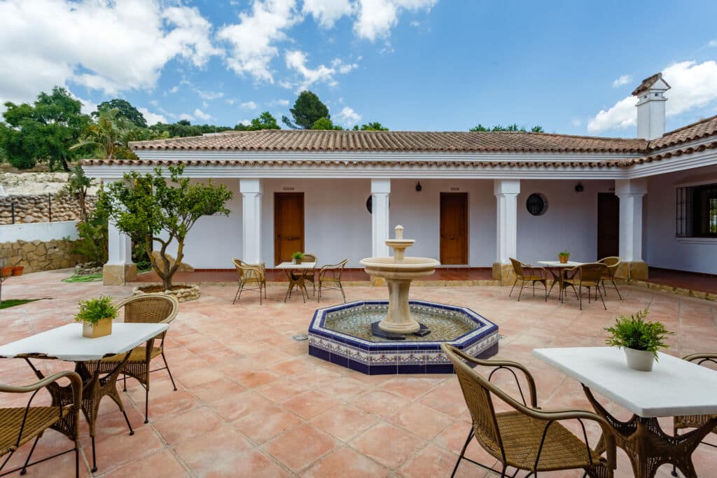 Un patio soleado con una fuente central de piedra, rodeado de mesas y sillas de mimbre. En las mesas hay macetas con plantas y, al fondo, columnas blancas con puertas de madera bajo un cielo azul.