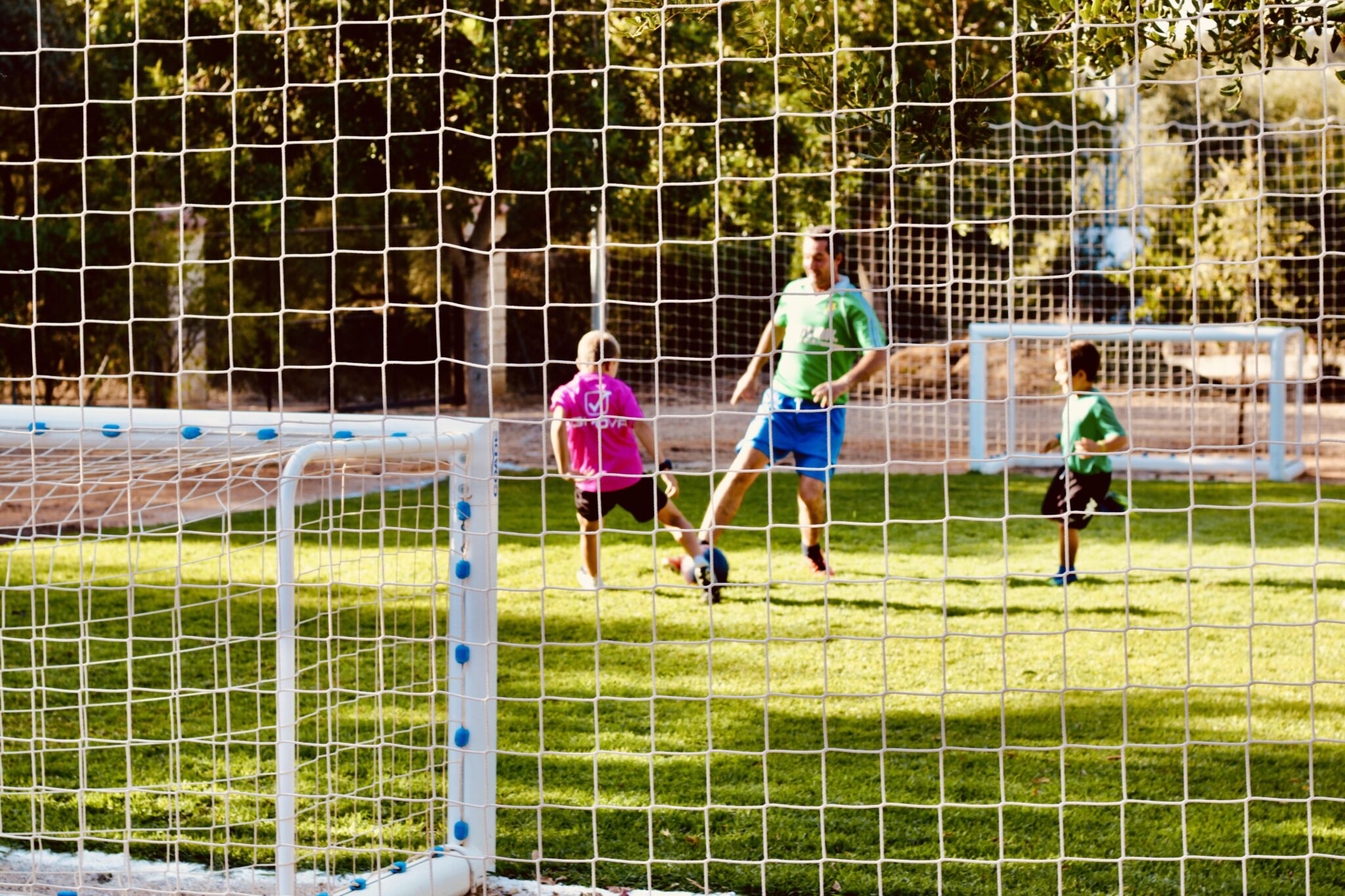 Tres personas, dos niños y un adulto, juegan al fútbol en un campo de hierba. La foto está tomada desde detrás de una red de portería, con árboles y la luz del sol de fondo. Los niños llevan camisetas rosas y verdes.
