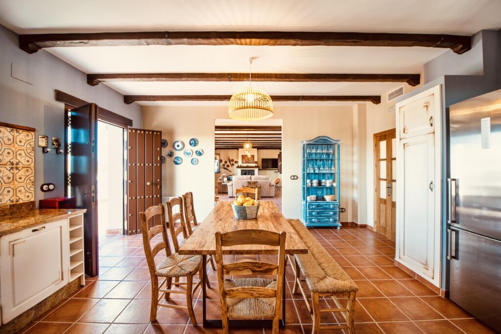 Una cocina y un comedor rústicos con una mesa de madera, seis sillas, vigas vistas en el techo, suelo de baldosas, un armario de porcelana azul, una lámpara colgante y puertas dobles abiertas que dejan pasar la luz natural.