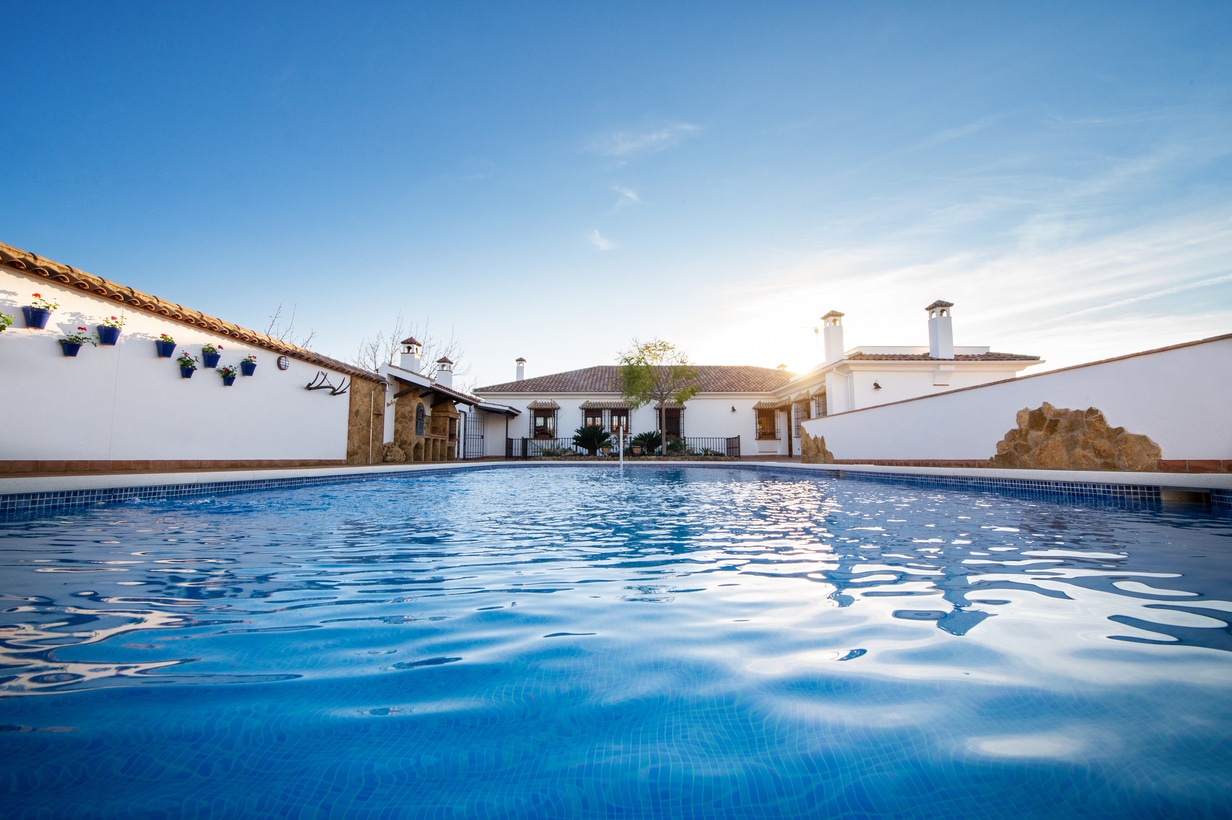 Vista desde un ángulo bajo de una piscina de color azul claro rodeada de edificios de paredes blancas con tejados de tejas, plantas de exterior y la brillante luz del sol de fondo.