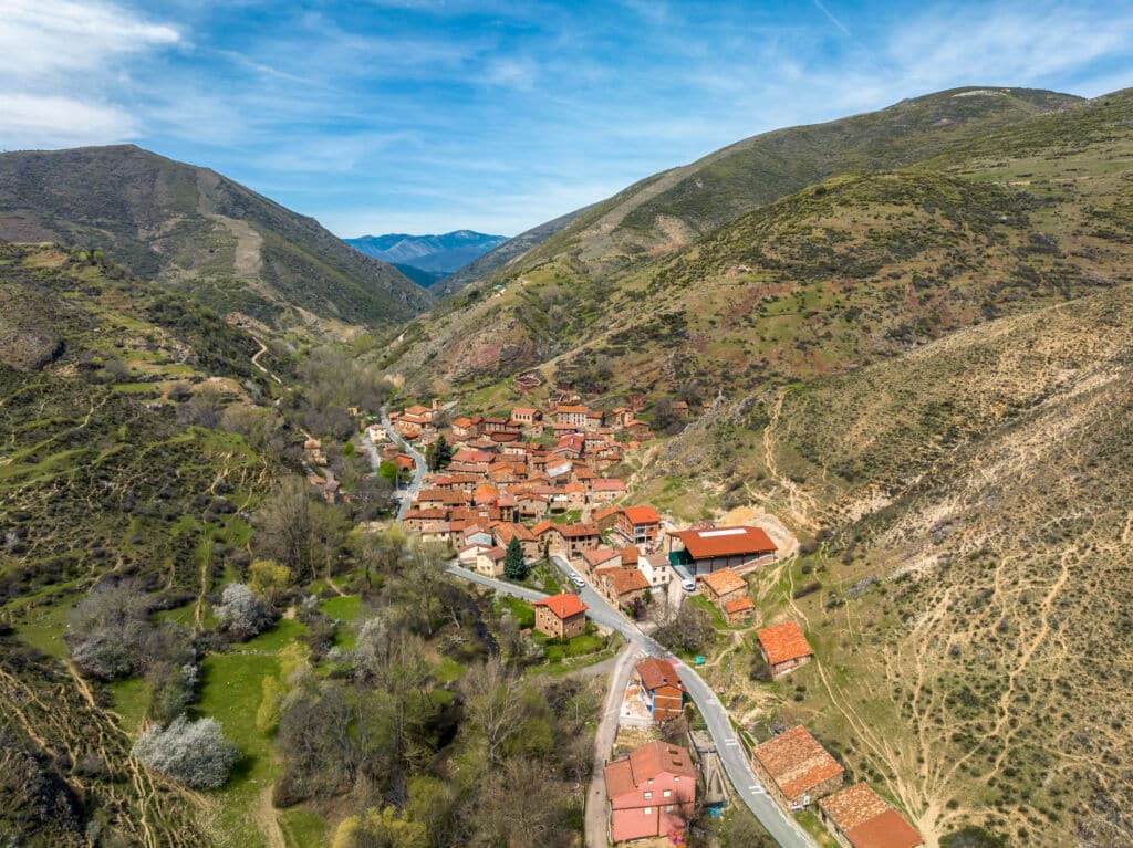 Vista aérea de un pequeño pueblo con tejados de tejas rojas enclavado en un valle verde entre colinas onduladas y montañas en un día claro y soleado. Una carretera serpenteante atraviesa el pueblo y el paisaje circundante.
