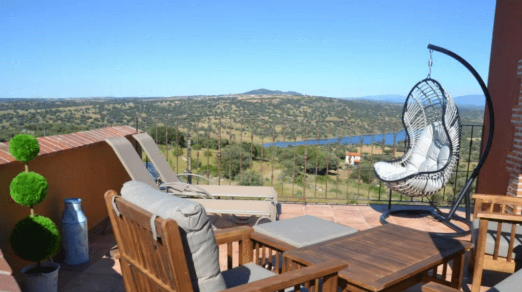 Una soleada terraza con sillas de madera, una mesa, dos tumbonas y una silla huevo colgante ofrece vistas a un pintoresco paisaje de verdes colinas, árboles y un río bajo un cielo azul despejado.