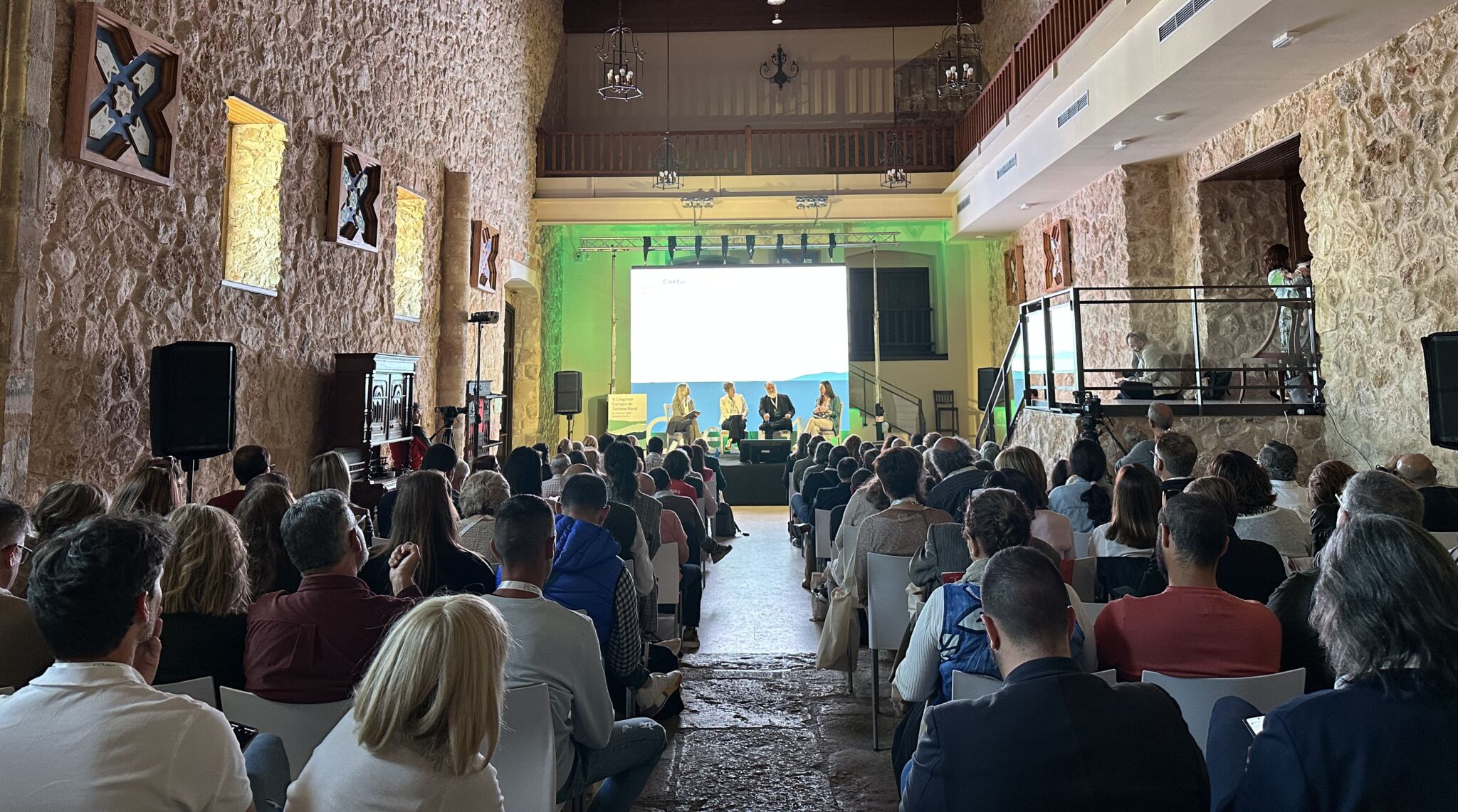 Un numeroso público se sienta en filas frente a un escenario muy iluminado con un panel de oradores en una sala rústica con paredes de piedra, techos altos y balcones. El acto parece ser una conferencia o un seminario.