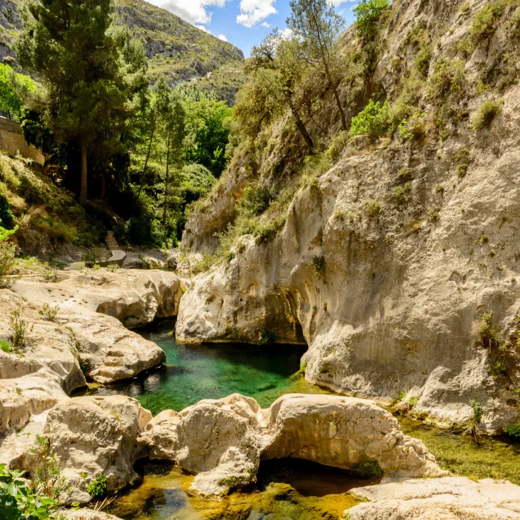 Una piscina cristalina de color verde esmeralda enclavada entre escarpadas rocas calizas y acantilados, perfecta para una escapada rural. Rodeada de árboles y exuberante vegetación bajo un cielo parcialmente nublado, invita a vivir el auténtico turismo rural.