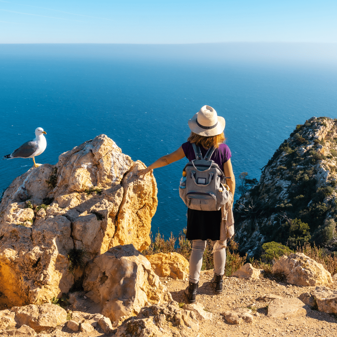 Una mujer con sombrero y mochila se encuentra en un acantilado rocoso con vistas al océano durante un verano rural en la Comunitat Valenciana, extendiendo la mano hacia una gaviota posada cerca, con el cielo azul claro y el agua lejana de fondo.