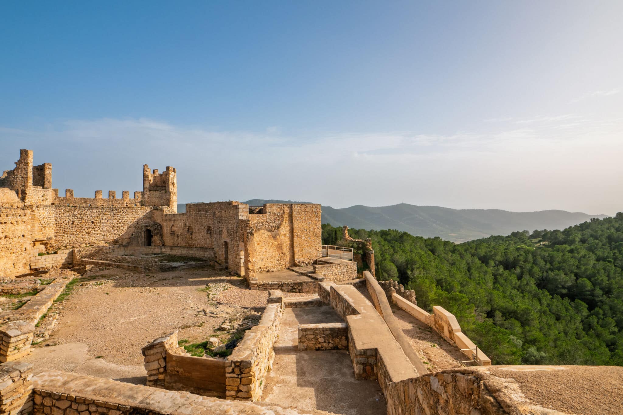 Las ruinas de una antigua fortaleza de piedra con altas murallas y torres se asoman a un frondoso bosque verde y a montañas lejanas bajo un cielo azul despejado, capturando el encanto de un verano rural en la Comunitat Valenciana.