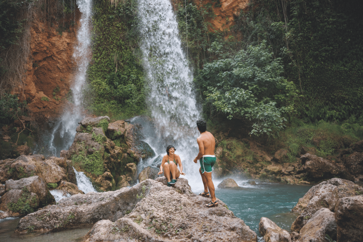 Dos personas en bañador disfrutan de un verano rural en la Comunitat Valenciana, relajándose en las rocas cerca de una exuberante cascada: una sentada y la otra de pie junto al agua azul claro de la base de la cascada.