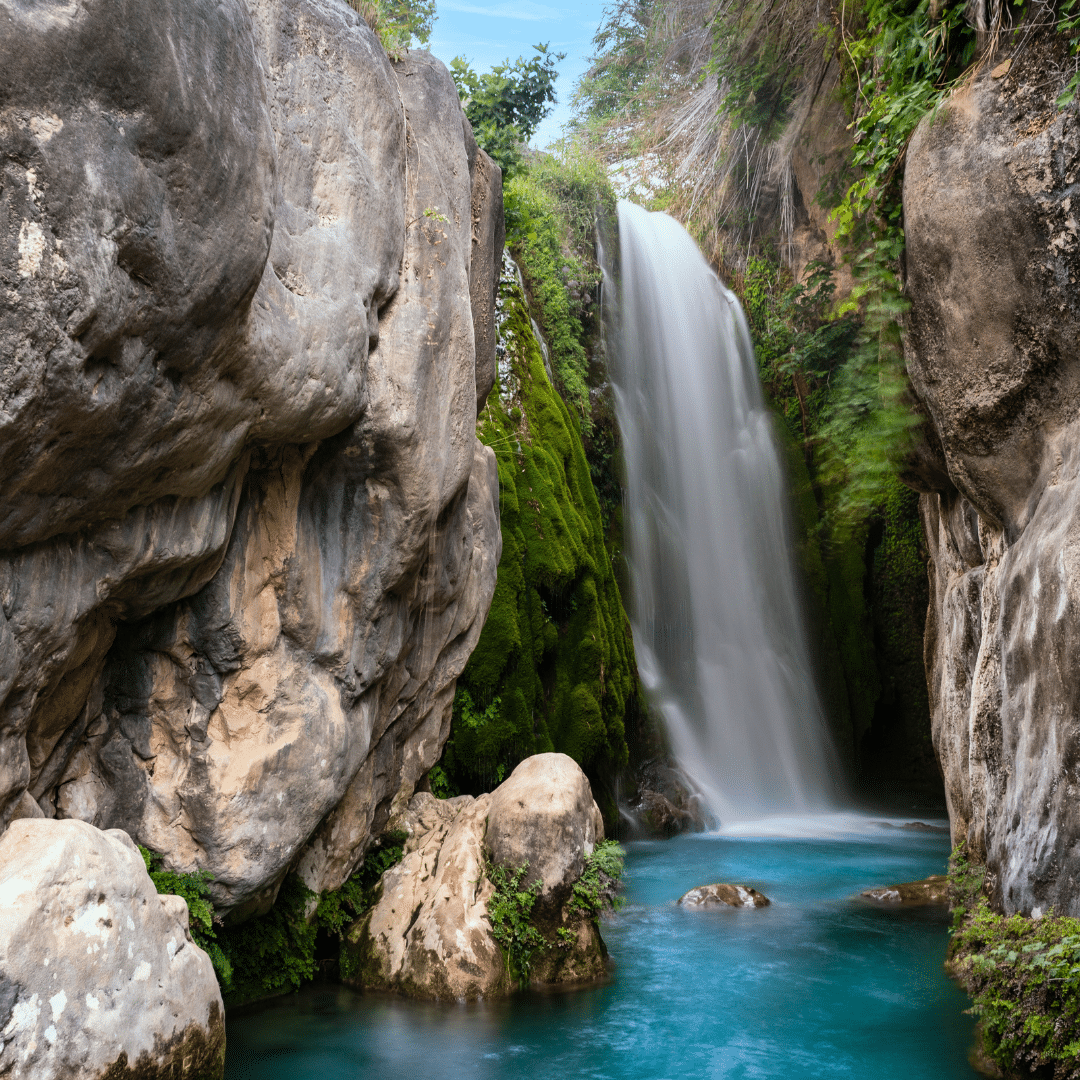 Una alta cascada cae entre rocas cubiertas de musgo a un estanque azul y transparente rodeado de exuberante vegetación y grandes rocas de piedra: una idílica escena de verano rural en la Comunitat Valenciana bajo un cielo parcialmente nublado.