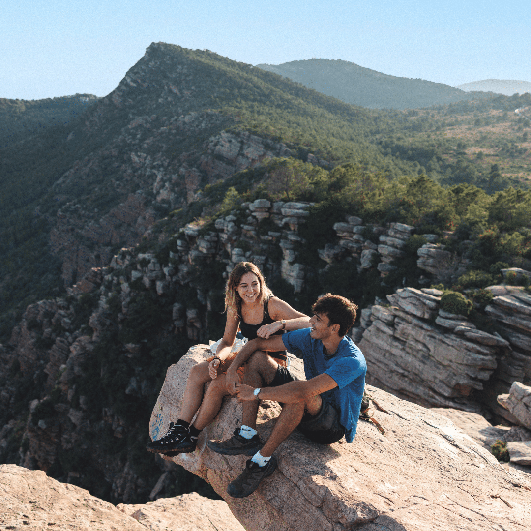 Dos personas sentadas al borde de un acantilado rocoso, sonríen y conversan durante un verano rural en la Comunitat Valenciana, con vastos paisajes montañosos y verdes bosques que se extienden a sus espaldas bajo un cielo azul despejado.