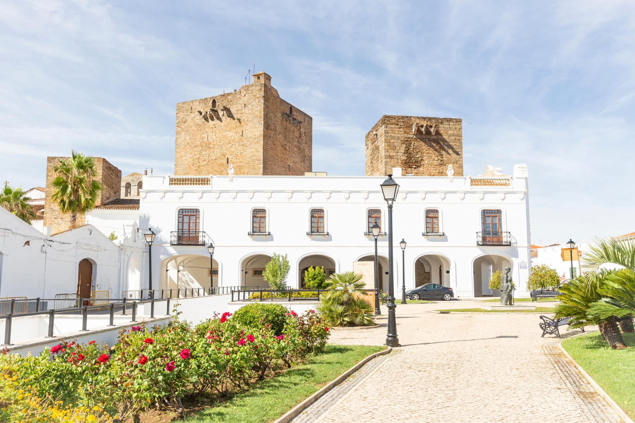 Edificio blanco con arcos y dos grandes torres de piedra detrás, rodeado de jardines, palmeras y un camino empedrado bajo un cielo azul. Un coche está aparcado cerca del edificio.