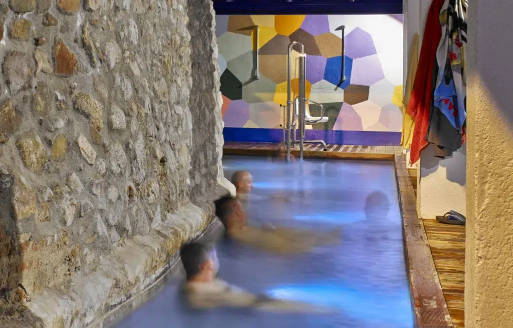Tres personas se relajan en una estrecha piscina cubierta junto a un muro de piedra: un lugar acogedor para una escapada rural. El fondo muestra un colorido mural geométrico, un elevador metálico y toallas colgadas en el suelo de madera.