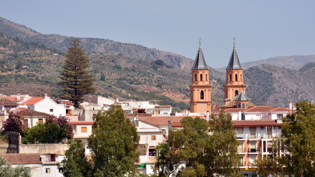 Vista panorámica de una ciudad con edificios con tejados de terracota, dos torres de iglesia con chapiteles y árboles verdes en primer plano, con un telón de fondo de colinas onduladas y montañas bajo un cielo despejado.