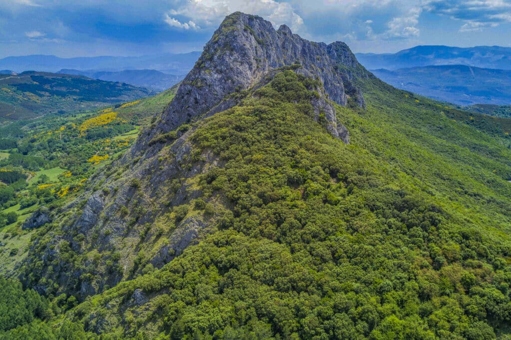 Una cima rocosa y escarpada, cubierta de un denso bosque verde, se eleva abruptamente contra un cielo azul -un lugar ideal para una escapada rural- rodeada de colinas onduladas y montañas lejanas bajo nubes dispersas.