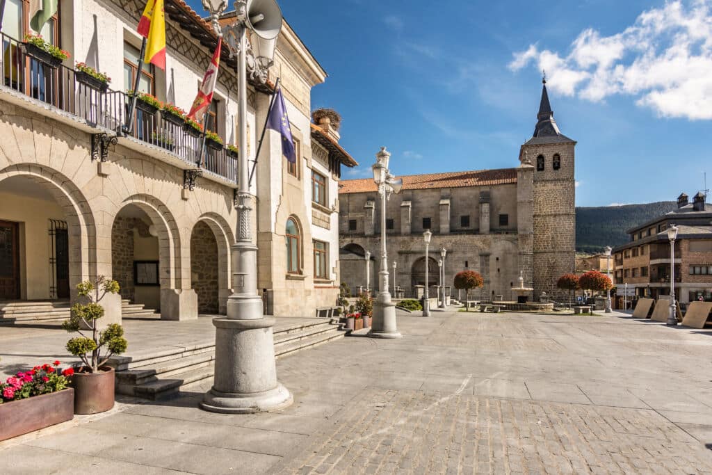 Una soleada plaza con edificios de piedra, pasarelas arqueadas, macetas con flores, balcones adornados con banderas y una iglesia con un alto campanario bajo un cielo azul con nubes dispersas.