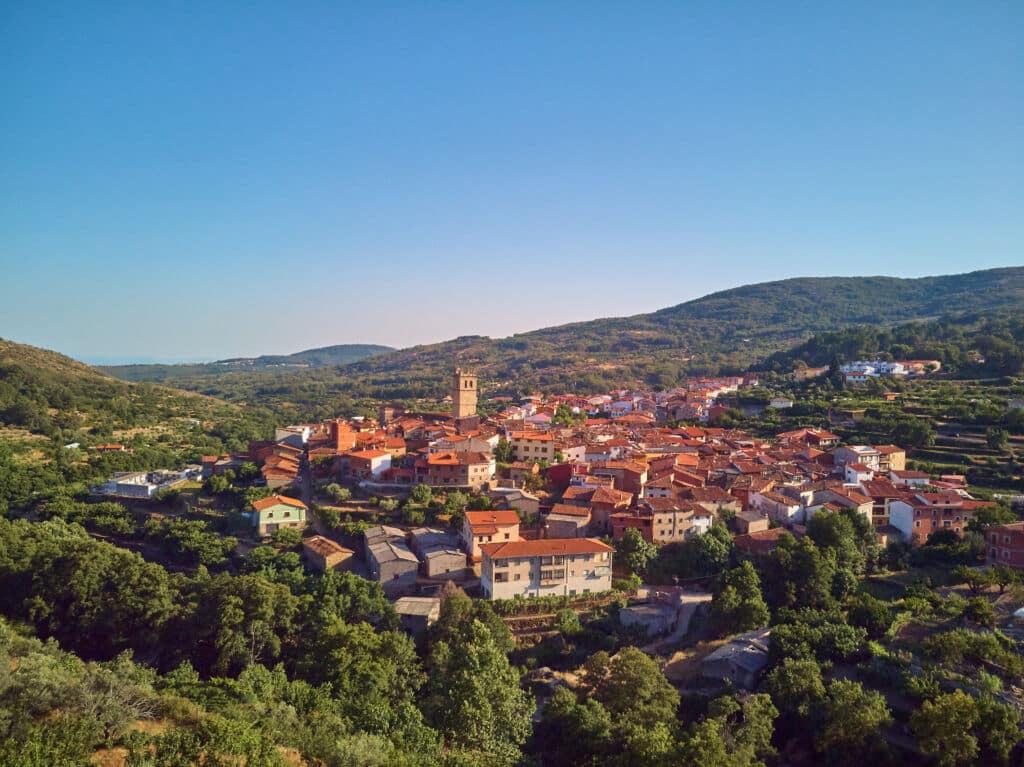 Vista aérea de un pequeño pueblo con casas de tejados rojos y una prominente torre de iglesia, rodeado de verdes colinas y árboles bajo un cielo azul despejado.