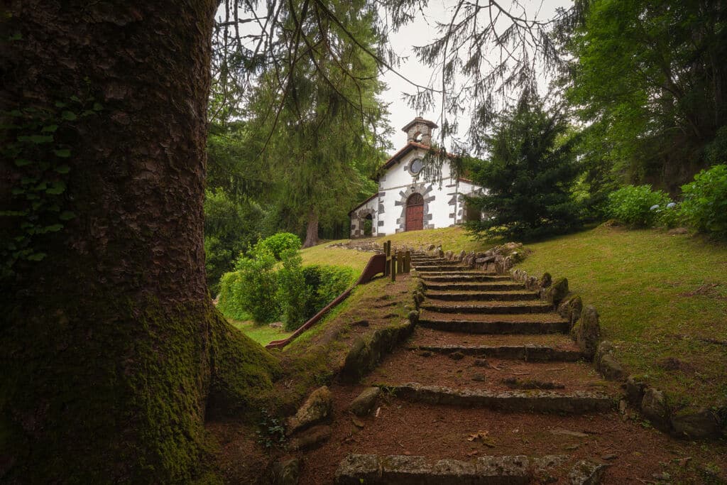 Unos escalones de piedra suben por una colina verde y arbolada hacia una pequeña capilla blanca con una puerta de madera y una cruz en el tejado. La escena es apacible, rodeada de frondosa hierba y grandes árboles.