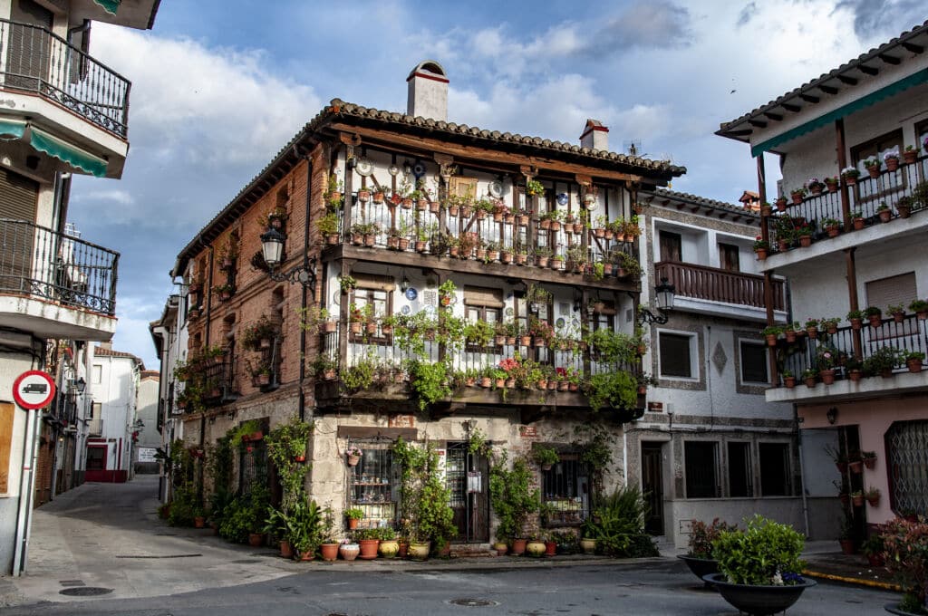Un encantador edificio rústico con balcones cubiertos de macetas y flores se alza en una tranquila esquina, rodeado de casas similares de estilo mediterráneo, bajo un cielo parcialmente nublado.