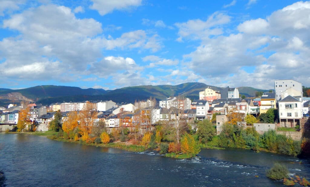 Una ciudad ribereña con coloridos árboles otoñales, edificios modernos y tradicionales, y montañas al fondo bajo un cielo azul con nubes dispersas.