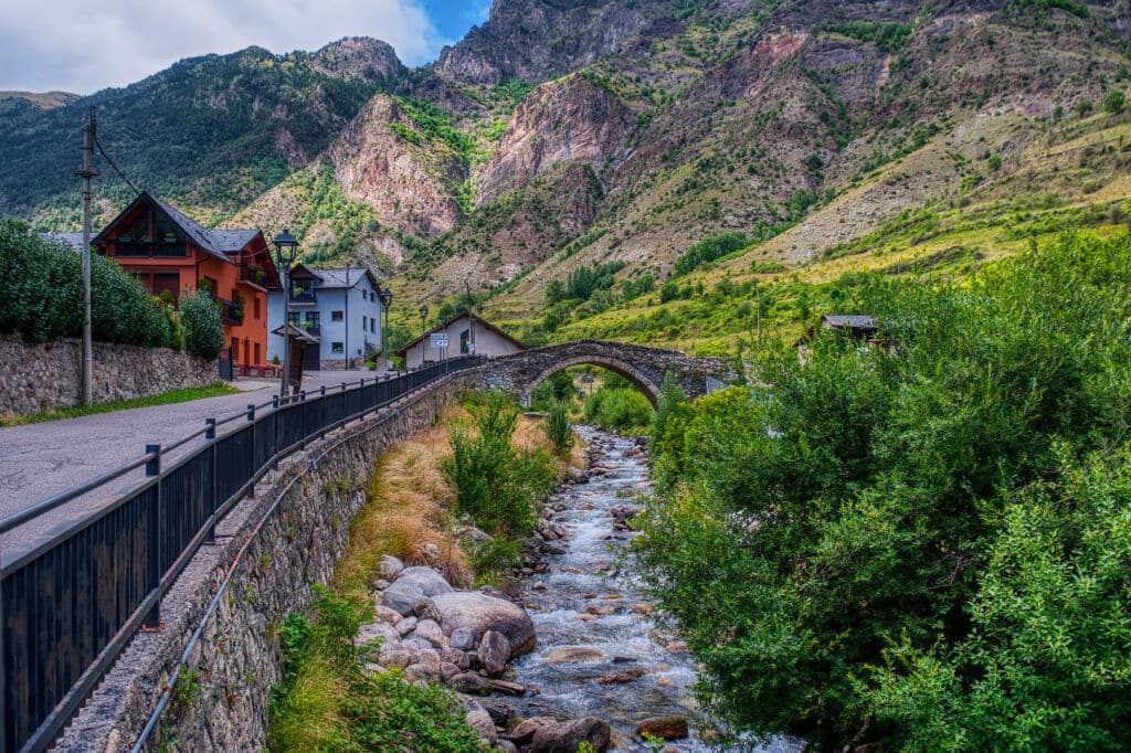 Un puente de piedra cruza un arroyo rocoso junto a casas de colores, con exuberantes montañas verdes elevándose al fondo bajo un cielo parcialmente nublado.