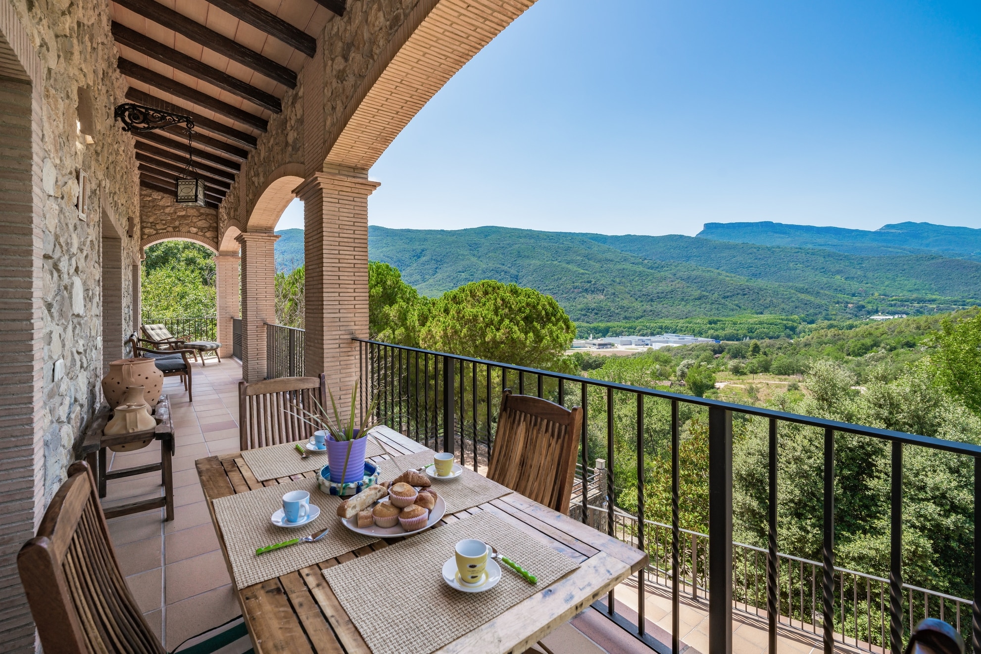 Una terraza de piedra cubierta con una mesa preparada para el desayuno ofrece vistas a exuberantes colinas verdes y a un amplio valle bajo un cielo azul despejado. Las sillas de madera y las plantas en macetas realzan la acogedora escena al aire libre.