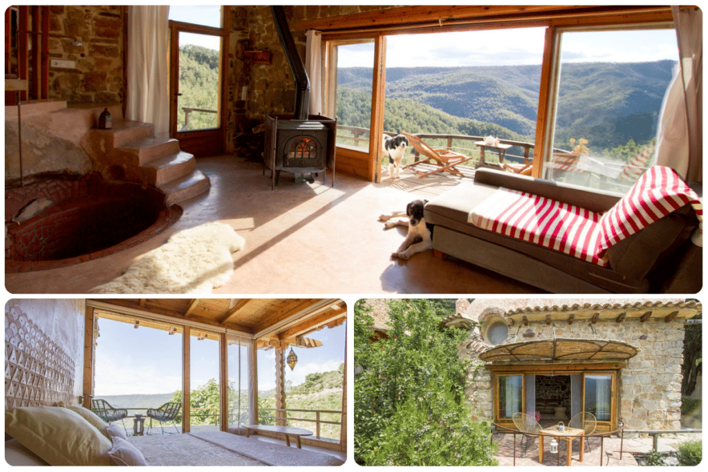 Una acogedora casa de piedra con grandes ventanales con vistas a las colinas boscosas. Dentro hay una bañera, una estufa de leña, un sofá y un perro. El patio exterior tiene una mesa y sillas, y un dormitorio se abre a la vista panorámica.