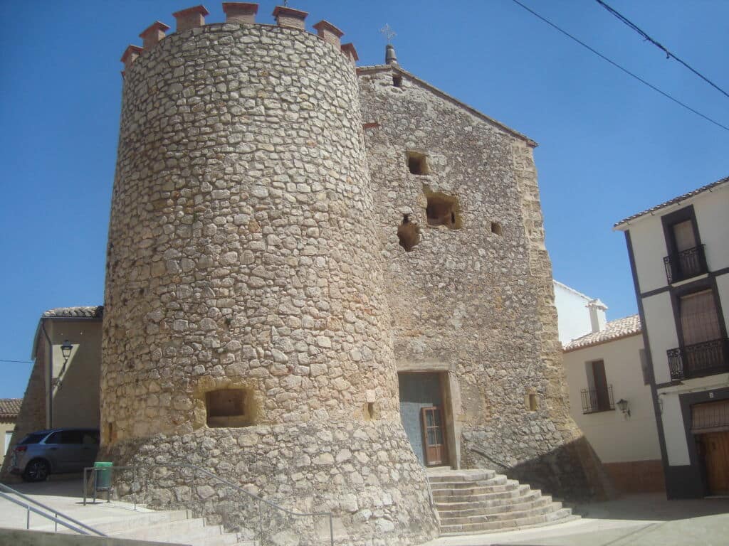 Una torre medieval de piedra con una torreta cilíndrica y pequeñas ventanas, adosada a un edificio con escaleras que conducen a una puerta de madera, rodeada de casas modernas bajo un cielo azul despejado.