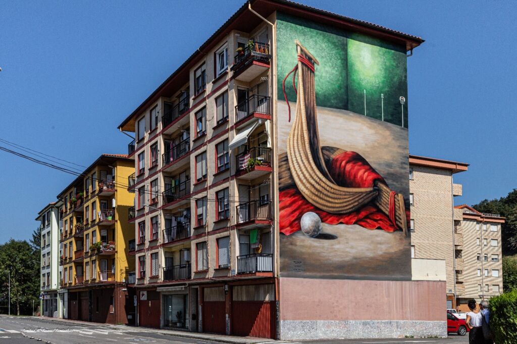 Un gran mural en el lateral de un edificio de apartamentos representa una figura surrealista hecha de formas alargadas y verticales envuelta en una tela roja, con una bola plateada en primer plano y un fondo verde y abstracto.