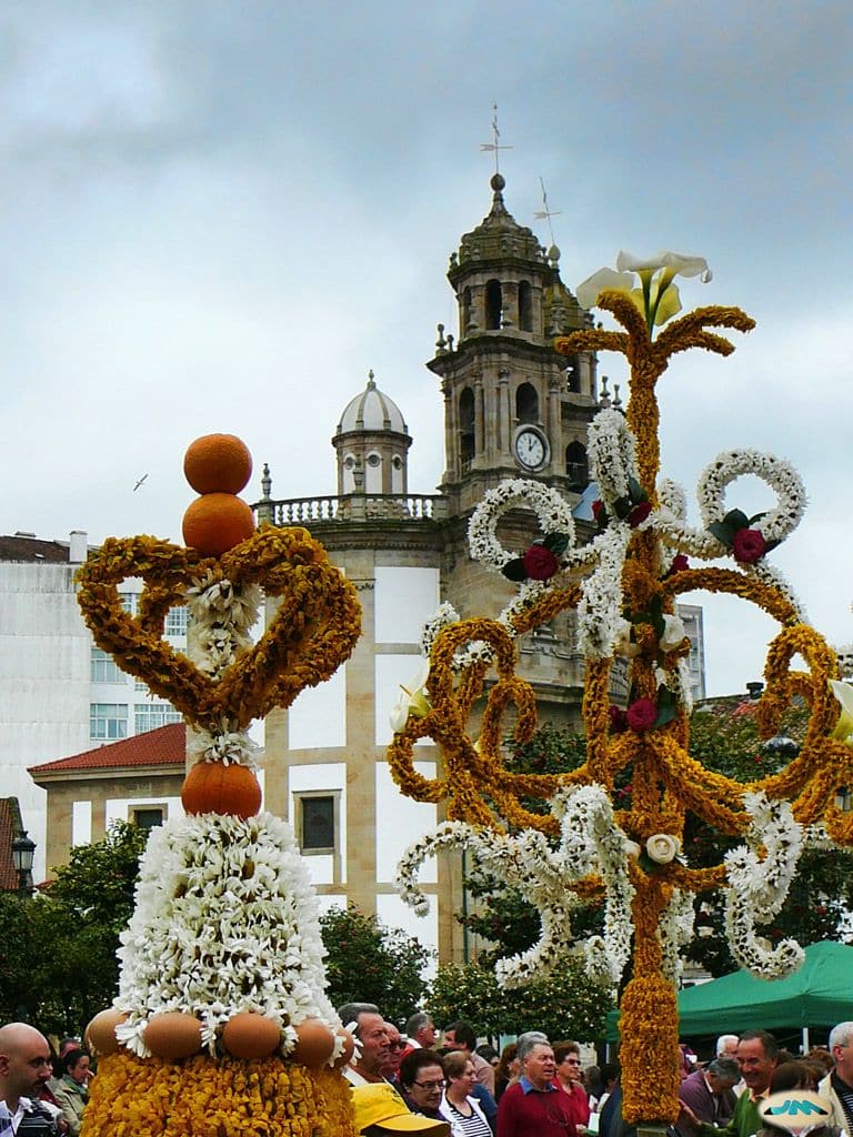 Estructuras florales decorativas adornadas con flores, frutas y huevos se exhiben en una concurrida plaza al aire libre, con una iglesia histórica y la torre del reloj visibles al fondo bajo un cielo nublado.
