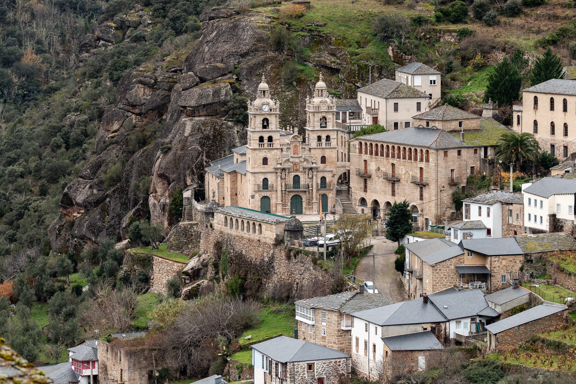 Edificios históricos de piedra y una gran iglesia con torres gemelas se asientan en una ladera rocosa rodeada de árboles y casas más pequeñas, confundiéndose con el paisaje montañoso.