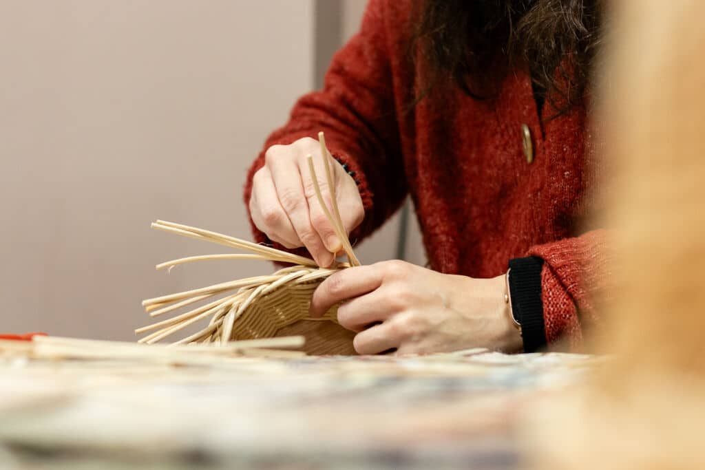Una persona vestida con un jersey rojo está tejiendo una cesta con juncos naturales o paja. Sus manos se concentran en el borde de la cesta, entrelazando cuidadosamente el material. El fondo está difuminado.