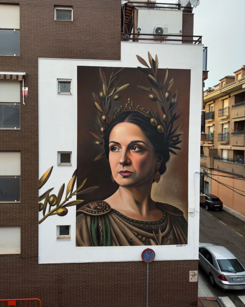 Un gran mural en un edificio muestra a una mujer regia con ramas de olivo a modo de corona y vestida con ropajes ornamentados. La pintura es de estilo clásico. En la calle se ven coches y edificios de apartamentos.
