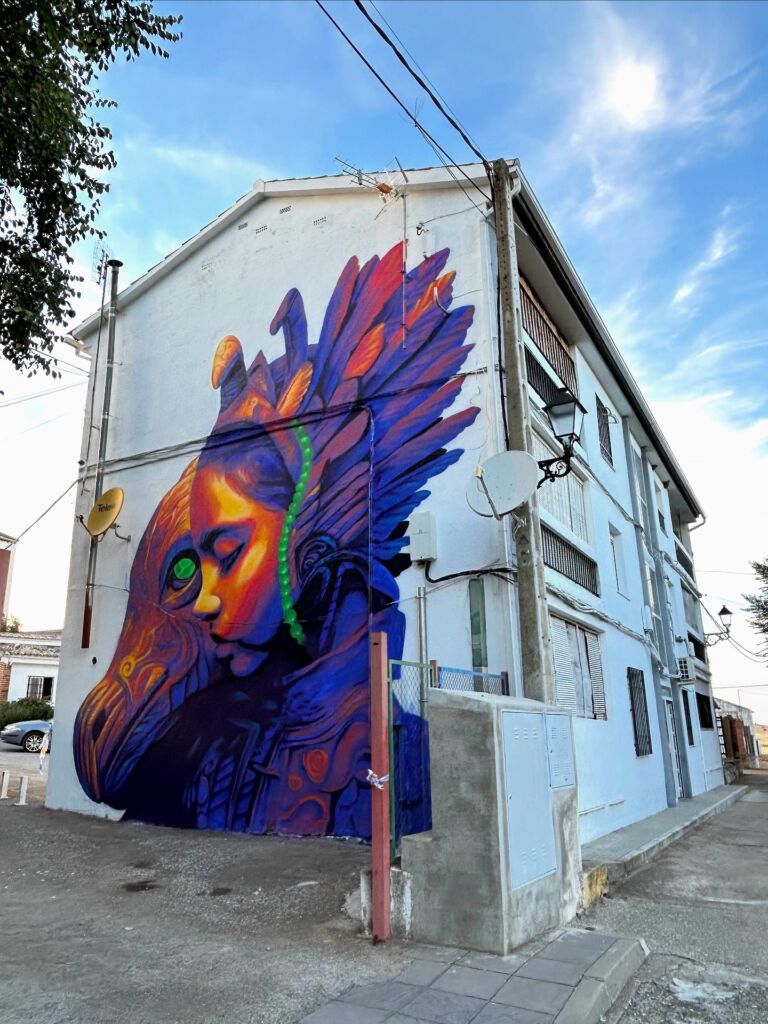 Un vibrante mural sobre un edificio blanco muestra un rostro humano que se funde con un colorido pájaro futurista, con vivos azules, morados, naranjas y verdes bajo un cielo brillante.