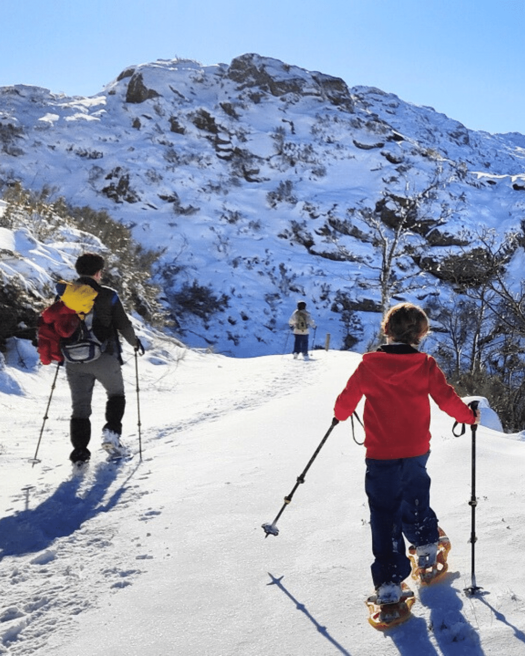 Tres personas, entre ellas un niño con chaqueta roja, caminan por un sendero de montaña nevado con bastones de trekking y raquetas de nieve, rodeados de rocas cubiertas de nieve y un cielo azul despejado.
