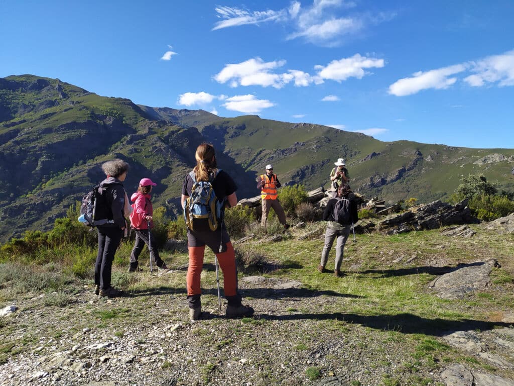 Un grupo de excursionistas con mochilas se encuentra en la cima de una colina cubierta de hierba, frente a un guía, con un telón de fondo de verdes montañas y un cielo azul brillante con nubes dispersas.