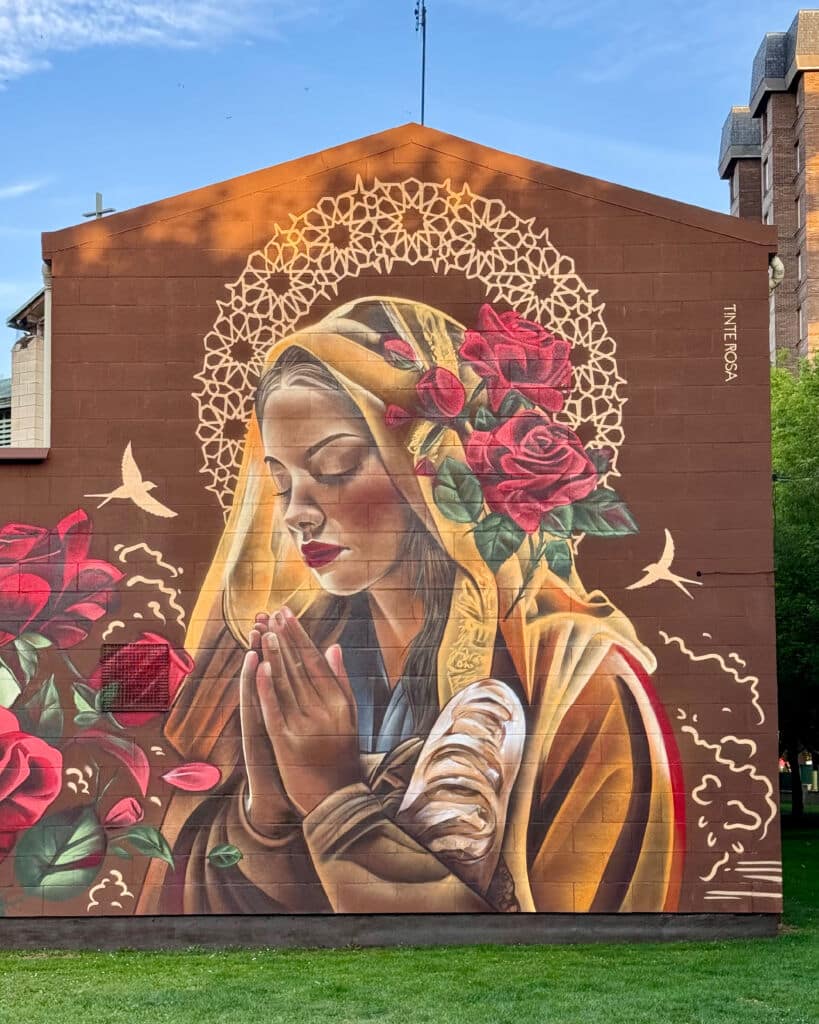 Un vibrante mural sobre un edificio muestra a una mujer con un chal y las manos en oración, rodeada de rosas, pájaros y un halo dorado sobre su cabeza. El fondo es de un intenso color marrón, con hierba verde en primer plano.