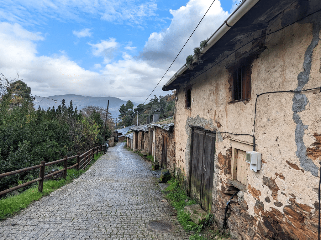 Una estrecha calle adoquinada bordeada de casas rústicas de piedra desgastada bajo un cielo parcialmente nublado, con árboles y montañas al fondo.