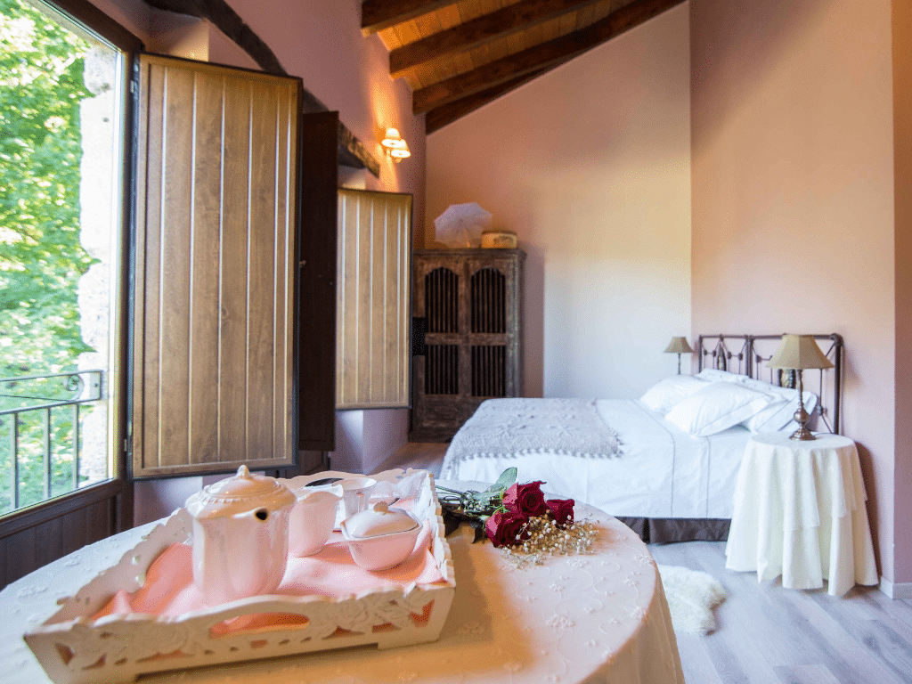 Un acogedor dormitorio con cama de matrimonio, techo de madera y ventanas abiertas con postigos. Una mesa en primer plano sostiene una bandeja con una tetera, tazas y rosas rojas. La cálida luz del sol ilumina la habitación.