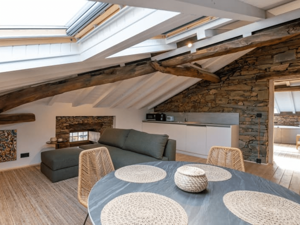 Un acogedor salón abuhardillado con vigas de madera vistas, paredes de piedra, un sofá verde, una mesa de comedor redonda con manteles individuales tejidos y una claraboya que deja pasar la luz natural. La cocina es compacta, con armarios blancos.