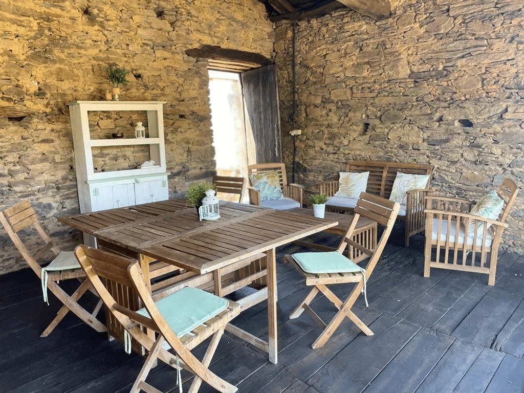 Habitación rústica con paredes de piedra, suelo de madera y muebles de madera que incluyen una mesa, sillas y asientos acolchados. En la pared hay un armario blanco con plantas y la luz del sol entra por una puerta abierta.