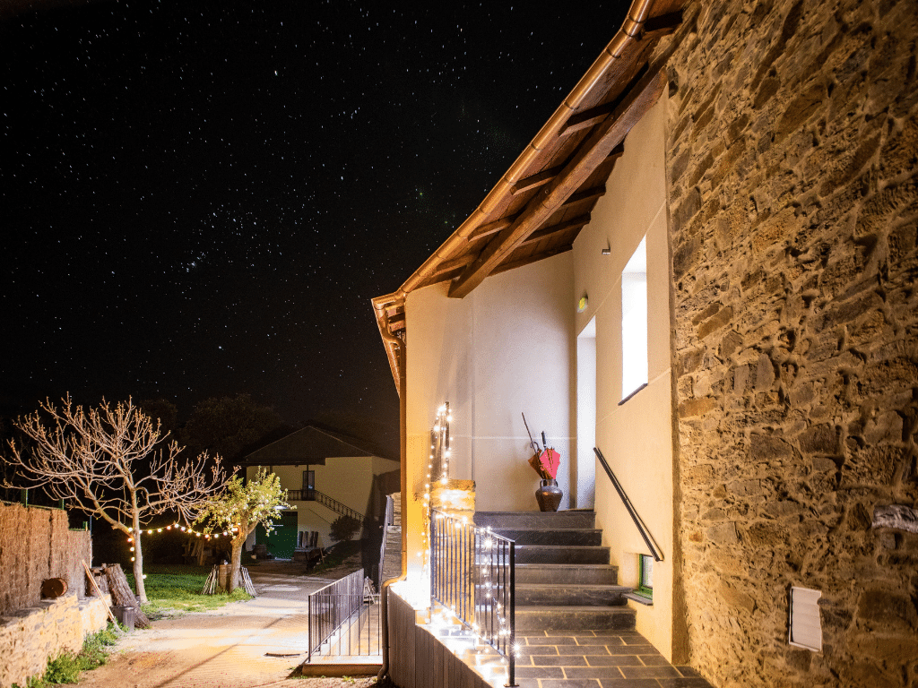 Una casa rural cálidamente iluminada con paredes de piedra y escaleras decoradas con cuerdas de luces, enmarcada en un cielo nocturno claro y estrellado. Al fondo se ven un árbol sin hojas y otro edificio.