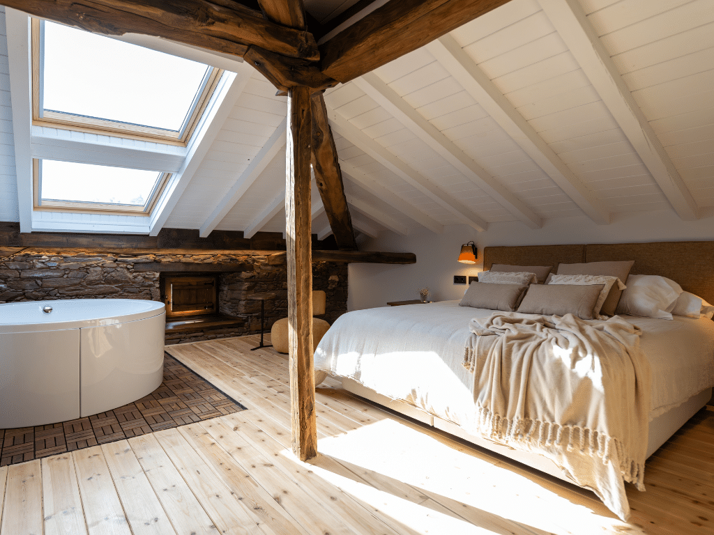 Un acogedor dormitorio abuhardillado con una cama grande, ropa de cama neutra, vigas de madera, una claraboya, una pared de piedra y una bañera exenta blanca cerca de una ventana. La cálida luz del sol inunda la habitación, creando un ambiente relajante.