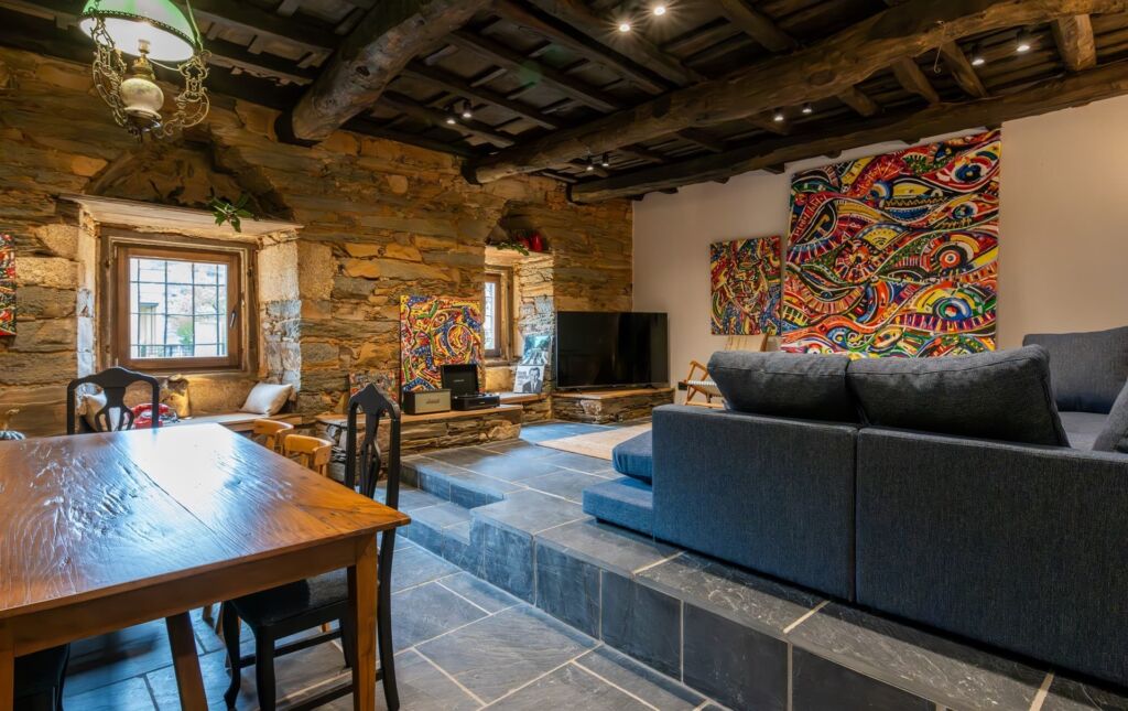 Una acogedora sala de estar con paredes de piedra, techo de madera oscura, coloridos cuadros abstractos, un sofá seccional gris, un televisor y una mesa de comedor de madera con sillas sobre un suelo de baldosas.