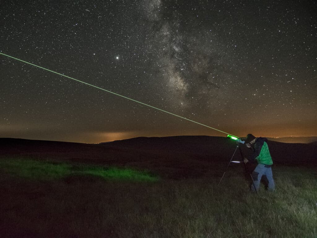 Una persona utiliza un puntero láser verde apuntando al cielo nocturno lleno de estrellas, con la Vía Láctea visible por encima de colinas oscuras y suelo cubierto de hierba, bajo un cielo claro y estrellado.
