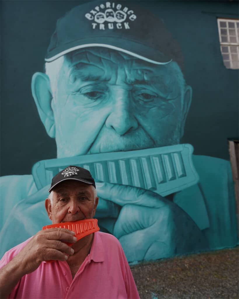 Un anciano con camisa rosa y gorra negra sostiene una armónica roja cerca de la boca, de pie frente a un gran mural en el que se representa a sí mismo tocando la armónica en tonos azules.